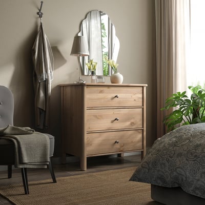 Chambre avec commode STOREMOLLA en bois clair, miroir, lampe et plante verte. Chaise et rideaux gris.