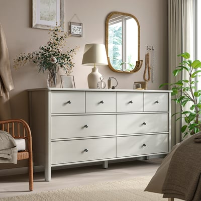Chambre moderne avec commode STOREMOLLA, lampe, miroir, fleurs. Chaise et lit en bois partiellement visibles.