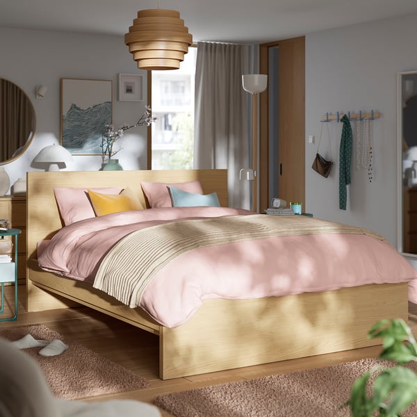 Chambre minimaliste avec lit en bois clair, literie rose, tapis et rideaux gris. Lit centré et simple flanqué d’armoires.