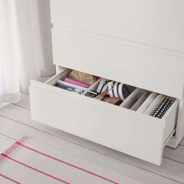 Commode blanche, ouverte pour montrer des vêtements pliés à l’intérieur, avec un tapis au sol.