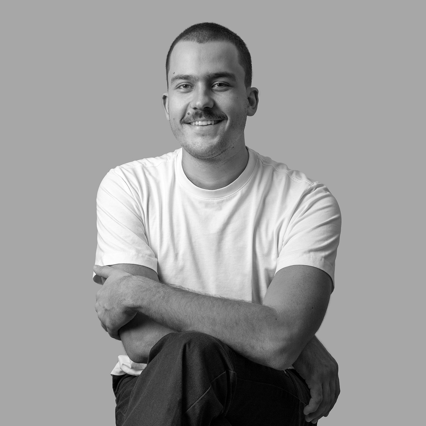 Photo en noir et blanc du créateur souriant assis, les bras croisés, portant un T-shirt clair et un jean foncé.