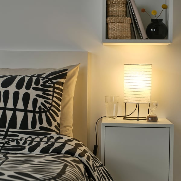Petite chambre avec literie noire et blanche, oreiller à motifs, table de chevet et lampe STRANDAD noire moderne. Les étagères au-dessus contiennent des paniers et un vase.