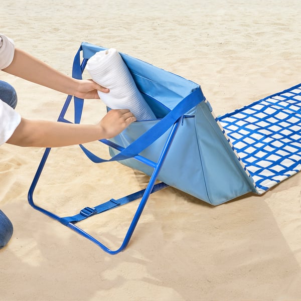Une personne place une serviette blanche dans un sac en tissu bleu pour chaises longues STRANDÖN avec des fermetures éclair sur une plage de sable.