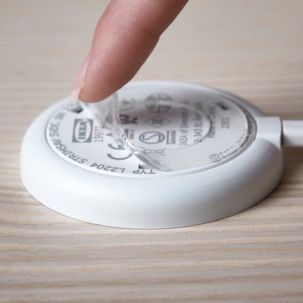 Une main appuyant un projecteur STRIMSÄV rond et blanc sur une surface en bois. Le projecteur s’allume lorsqu’il est pressé, adapté aux armoires et aux placards.