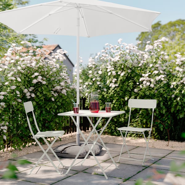 Petit patio avec table pliante blanche et chaises, boissons rouges, sous parasol blanc, entouré de buissons en fleurs.