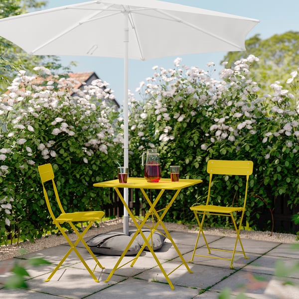 Table et chaises d’extérieur jaune vif avec parasol blanc, entourées de buissons en fleurs, idéales pour les petits espaces.