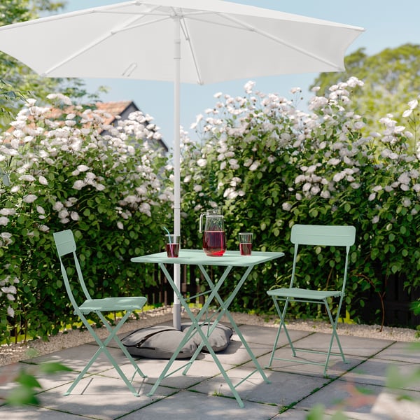 Scène de patio sereine avec table et chaises en métal SUNDSÖ vert clair, parasol blanc, fleurs roses et boissons.