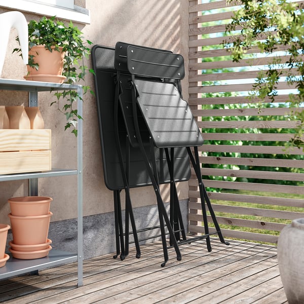 Trois chaises SUNDSÖ noires, pliables et empilables, reposent contre une clôture en bois sur un balcon, à côté d’une étagère avec des plantes en pot.
