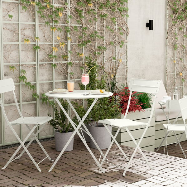 Petite installation extérieure avec table pliable blanche et chaises. Des plantes en pot entourent une salle à manger confortable.