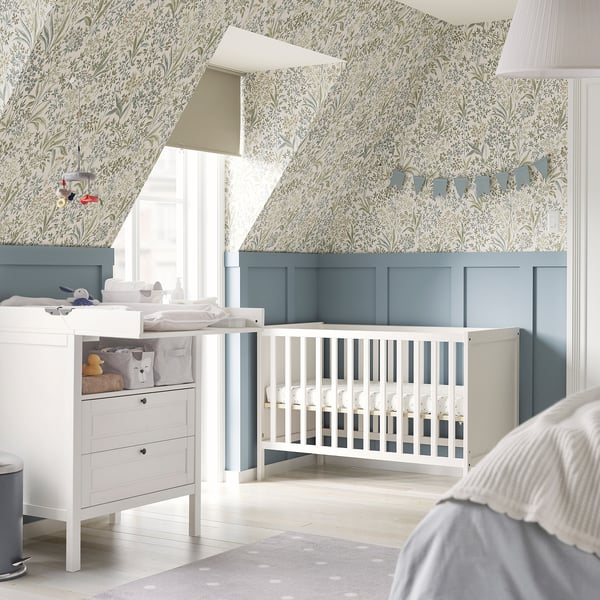 Chambre d’enfant avec lit SUNDVIK blanc, peinture bleue, papier peint floral, table à langer et tapis ; Décor doux et sûr.