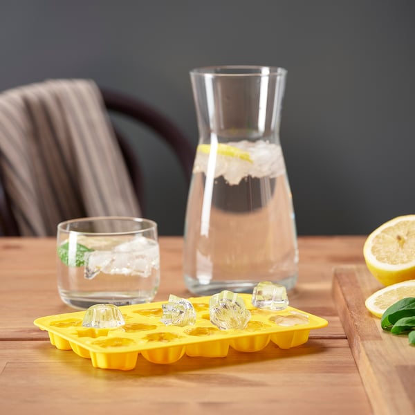 Bac à glaçons jaune vif avec des glaçons en forme de losange. Verre avec de la glace, une carafe d’eau et des citrons tranchés sur une table en bois.