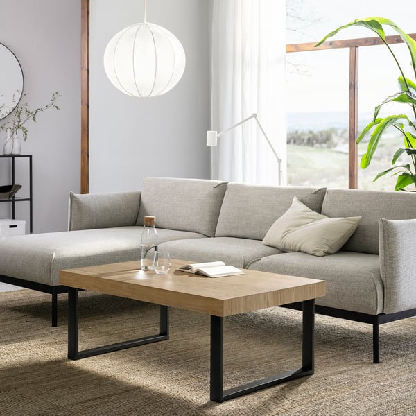 Grande table basse grise moderne avec pieds en métal et plateau en chêne, positionnée dans un salon avec un canapé gris, une suspension blanche et une plante verte.