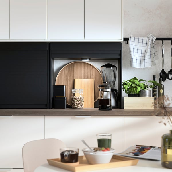 Cuisine moderne avec armoires TITTEBO noires, comptoir en bois et plantes. Chaises blanches et plateau avec boissons et fruits.