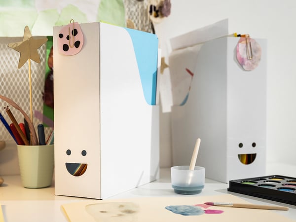 Deux classeurs de magazines TJABBA blancs et souriants, fabriqués à partir de carton ondulé 100 % recyclé, sur une table avec du matériel artistique.