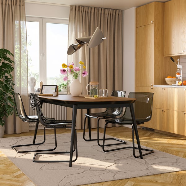 Salle à manger moderne avec table extensible en noyer, chaises noires et fleurs. Cuisine avec fenêtre et plante.