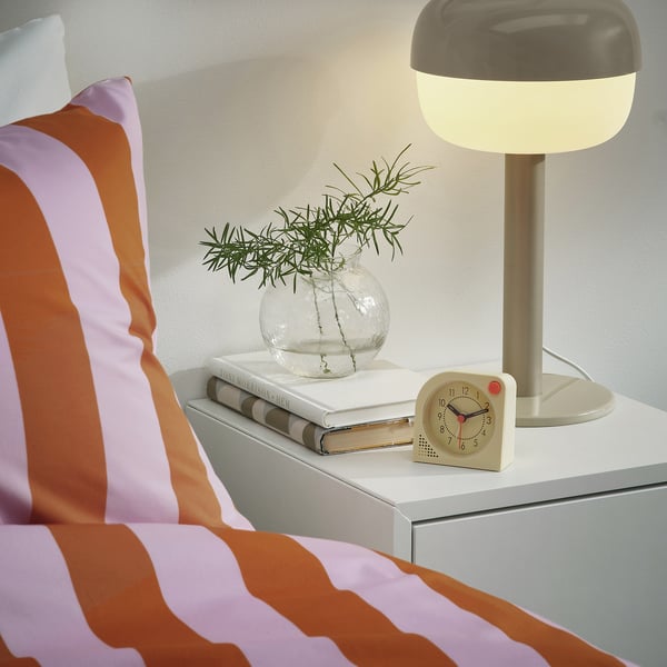 Lit rayé avec une table de chevet. La table contient des livres, un vase rond avec de la verdure, une horloge avec une alarme lumineuse et une lampe allumée.