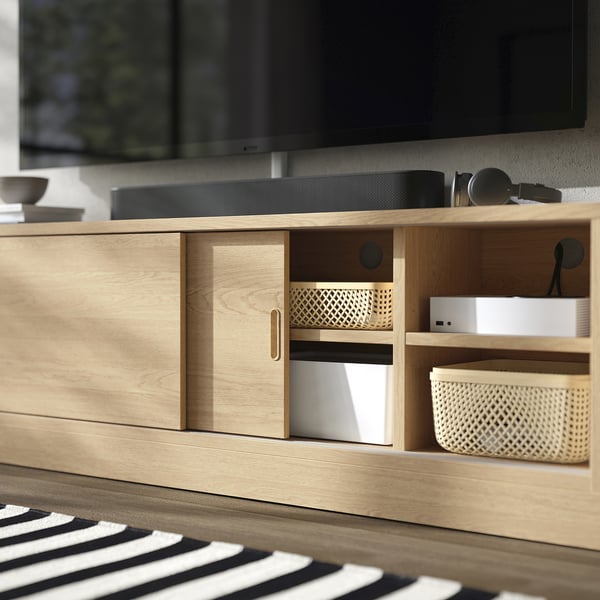 Meuble TV en bois avec portes coulissantes, gestion des câbles et étagères réglables, abritant des appareils électroniques et des bacs de rangement.