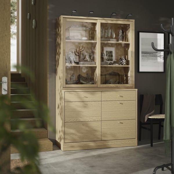 Une armoire en bois moderne nommée TONSTAD en finition chêne avec des portes en verre, exposant des objets décoratifs et six tiroirs pour le rangement.