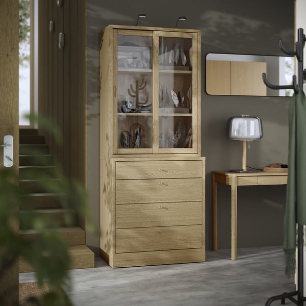 Armoire en bois brun clair avec portes et tiroirs en verre. Expose des objets décoratifs et organise le contenu. Un design épuré pour les espaces modernes.
