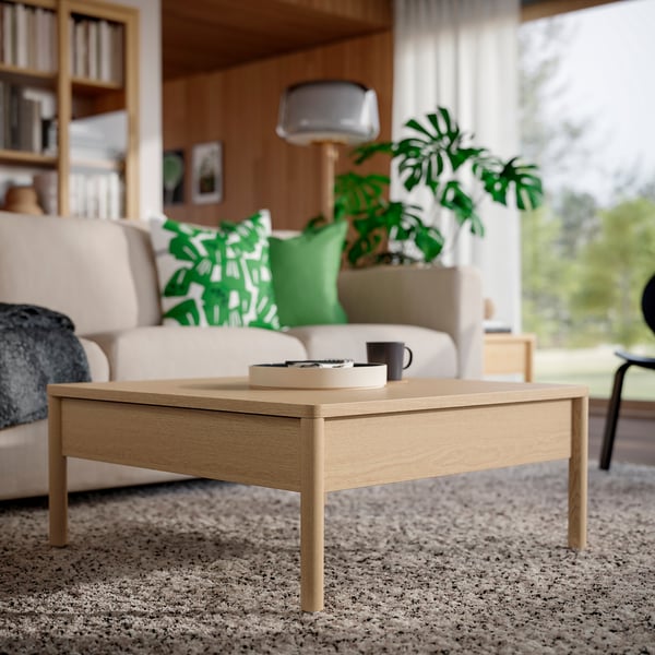Table basse en placage de chêne brun avec tiroirs latéraux sur moquette beige dans un salon confortable avec canapé, plante et étagère.
