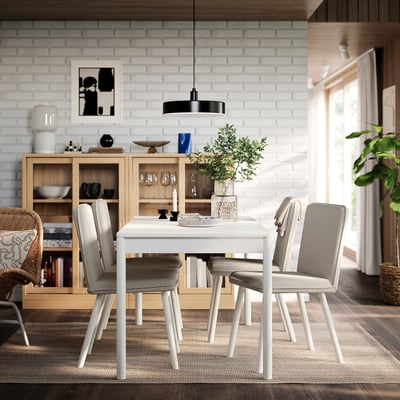 Un ensemble de salle à manger TONSTAD avec une table blanc cassé, des pieds en bois, quatre chaises, une étagère, un vase, des bougies et des œuvres d’art contre un mur de briques.