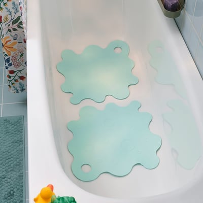 Deux tapis de bain TRAMPSJÖN turquoise clair avec ventouses dans une baignoire blanche à côté d’un rideau de douche coloré.