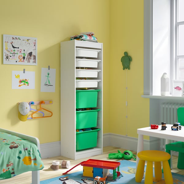 Chambre d’enfant lumineuse avec une grande unité de rangement TROFAST, un cadre blanc et des boîtes vertes, avec divers jouets et des œuvres d’art colorées sur des murs jaune clair.
