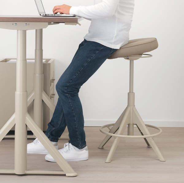 Personne assise sur un tabouret réglable, travaillant sur un ordinateur portable à un bureau réglable en hauteur.