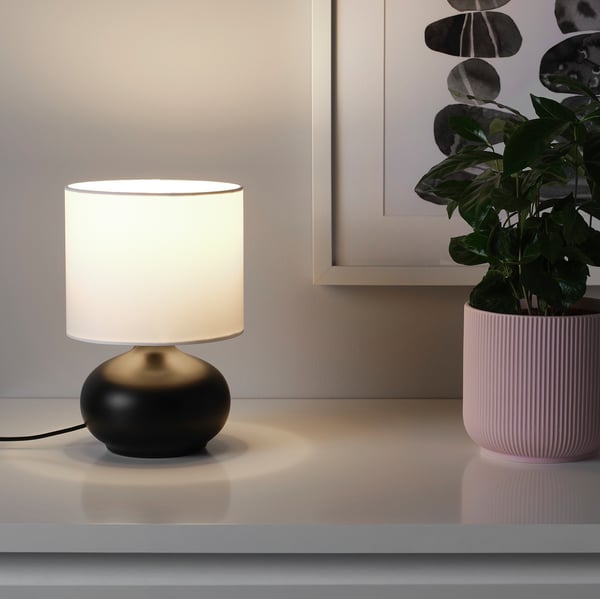 Une petite lampe de table avec une base en grès noir et un abat-jour blanc, émettant une lumière chaude.