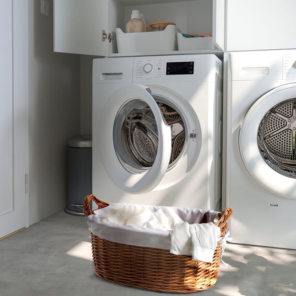 Une machine à laver blanche avec une porte ouverte, un panier à linge avec des draps à côté et des étagères de rangement au-dessus.