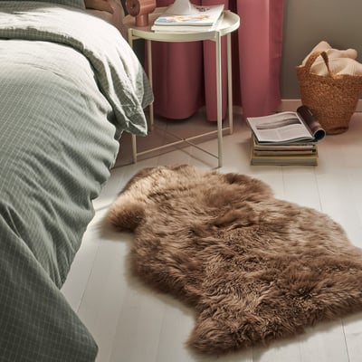 Chambre avec un lit vert, des rideaux roses et un tapis moelleux marron sur le sol. Une table d’appoint blanche contient des livres et une lampe. Un panier en osier se trouve à proximité.
