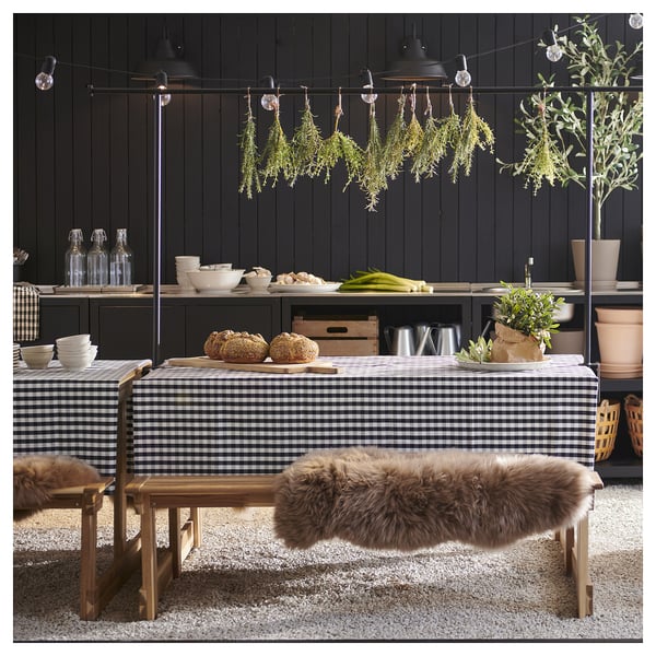 Salle à manger confortable avec nappe à carreaux noirs et blancs, table en bois, banc en peau de mouton, herbes suspendues et plantes en pot.