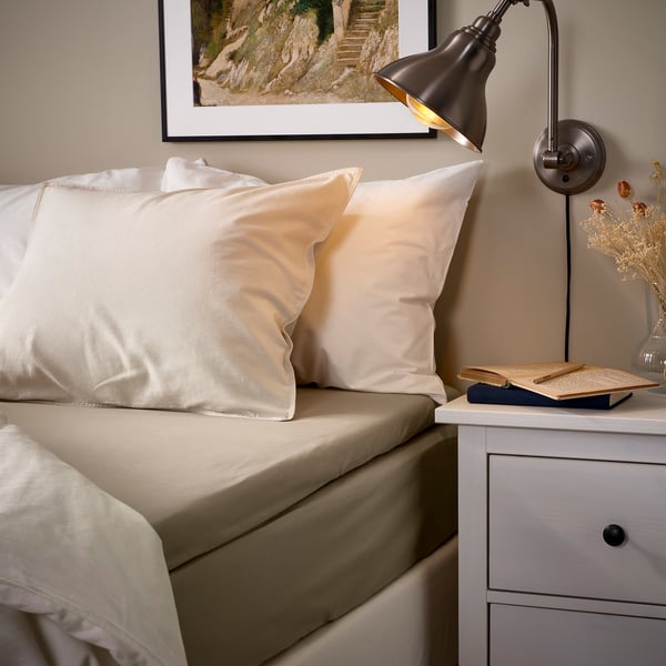 Lit douillet avec draps beiges ULLVIDE, lampe murale, œuvres d’art encadrées et vase de chevet. Tissu doux et respirant, oreillers légèrement plus grands.