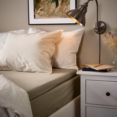 Lit douillet avec draps beiges ULLVIDE, lampe murale, œuvres d’art encadrées et vase de chevet. Tissu doux et respirant, oreillers légèrement plus grands.