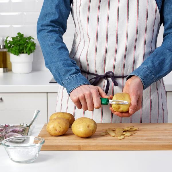 Personne épluchant des pommes de terre à l’aide d’un éplucheur à manche vert UPPFYLLD sur une planche de bois, entourée d’ingrédients de cuisine.