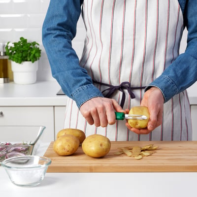 Personne épluchant des pommes de terre à l’aide d’un éplucheur à manche vert UPPFYLLD sur une planche de bois, entourée d’ingrédients de cuisine.