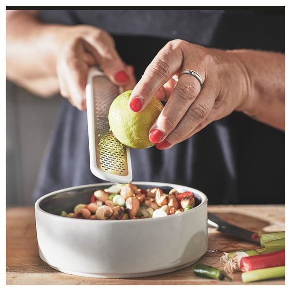 La personne râpe le zeste de citron vert dans un bol à l’aide d’une petite râpe à main UPPFYLLD blanche. Le bol contient un mélange de noix et de légumes hachés.