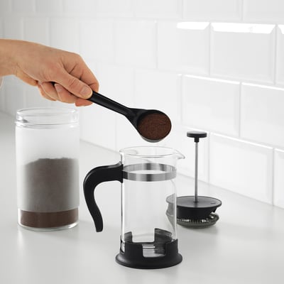 Une personne ajoute du marc de café à une presse française UPPHETTA transparente. Le piston est visible à côté, prêt à l’emploi sur un comptoir.