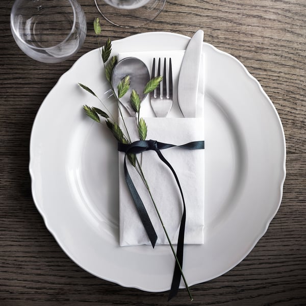Table à manger blanche avec assiette rustique UPPLAGA, bords à volants. Comprend une serviette de table, des ustensiles, de la verdure avec un ruban noir.