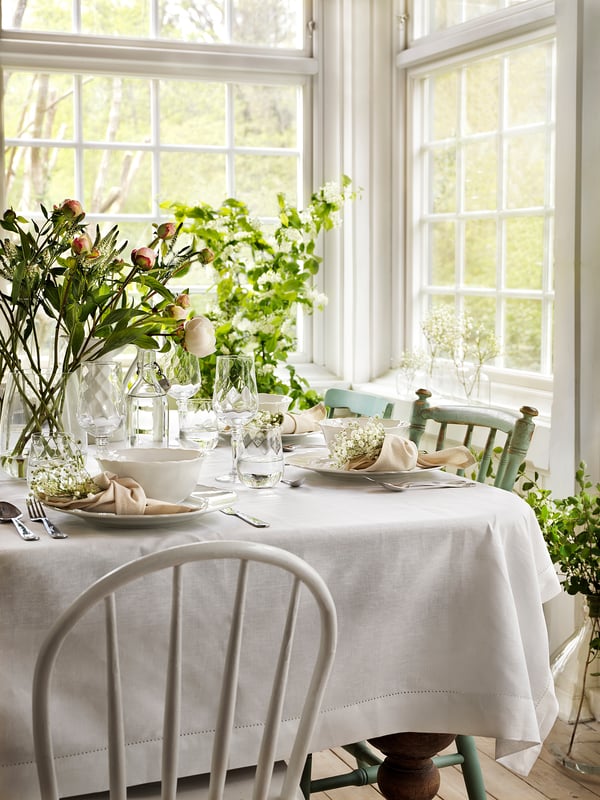 Salle à manger lumineuse avec vaisselle UPPLAGA blanche. Couverts et verrerie sur la table, entourés de centres de table floraux. De grandes fenêtres dévoilent de la verdure.