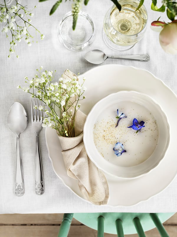 Bol de soupe blanc aux fleurs bleues. Cuillère et fourchette en argent, posées sur une serviette beige. Verres, vases, nappe légère. Porcelaine, design rustique.