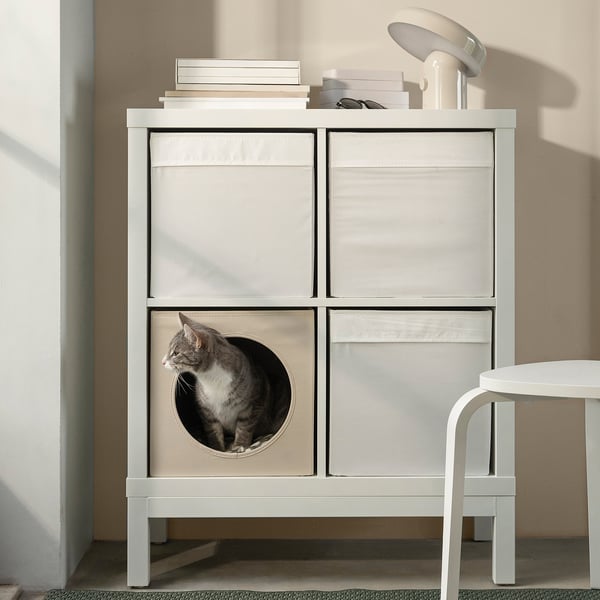 Niche à chat UTSÅDD blanche sur l’étagère Kallax, chat qui découle à travers une découpe circulaire. Livres et lampe dessus, tabouret à droite.