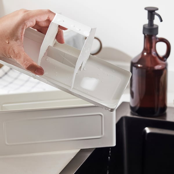 La main tient l’organisateur en plastique incliné, l’eau coule, bouteille brune avec pompe en arrière-plan. Garde la cuisine bien rangée.