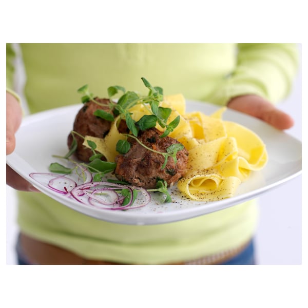 Personne tenant une assiette carrée blanche aux bords doux, avec de la viande grillée, de la sauce jaune, des pâtes et des oignons violets garnis d’herbes vertes.