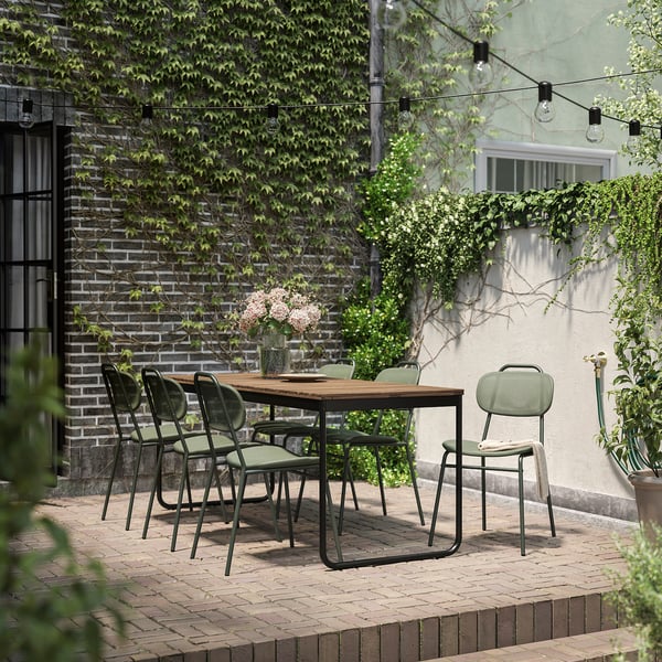 Salle à manger extérieure avec une table extensible en acacia et des chaises rembourrées vertes, entourée de briques et de verdure.