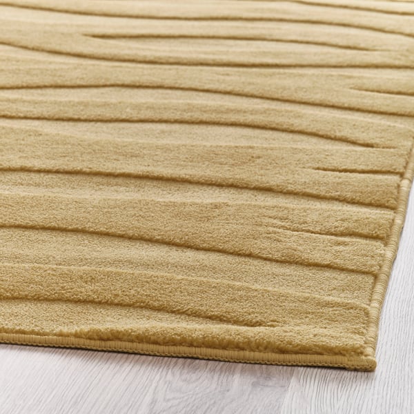 Tapis doux et ondulé jaune-brun avec un motif 3D texturé, posé sur un sol en bois clair.