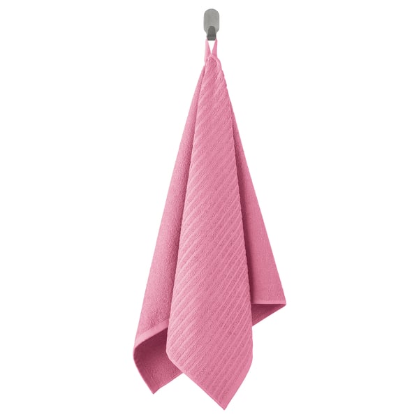 La serviette VÅGSJÖN rose vif est soigneusement suspendue à un crochet, avec un motif éponge doux et texturé composé à 100 % de coton.