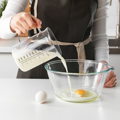 La personne verse le lait d’un pot doseur dans un bol avec un œuf. La verseuse a des marquages pour mesurer le liquide.