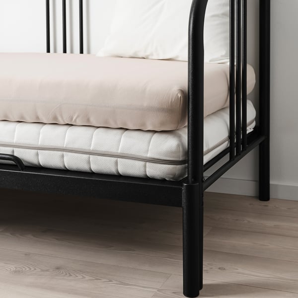 Lit en métal noir avec matelas beige et oreillers sur parquet.