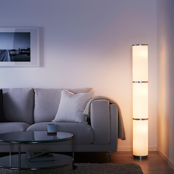 Salon moderne avec un grand lampadaire VIDJA fournissant une lumière ambiante chaleureuse à côté d’un canapé gris et d’une table basse en verre avec une tasse bleue et des magazines.